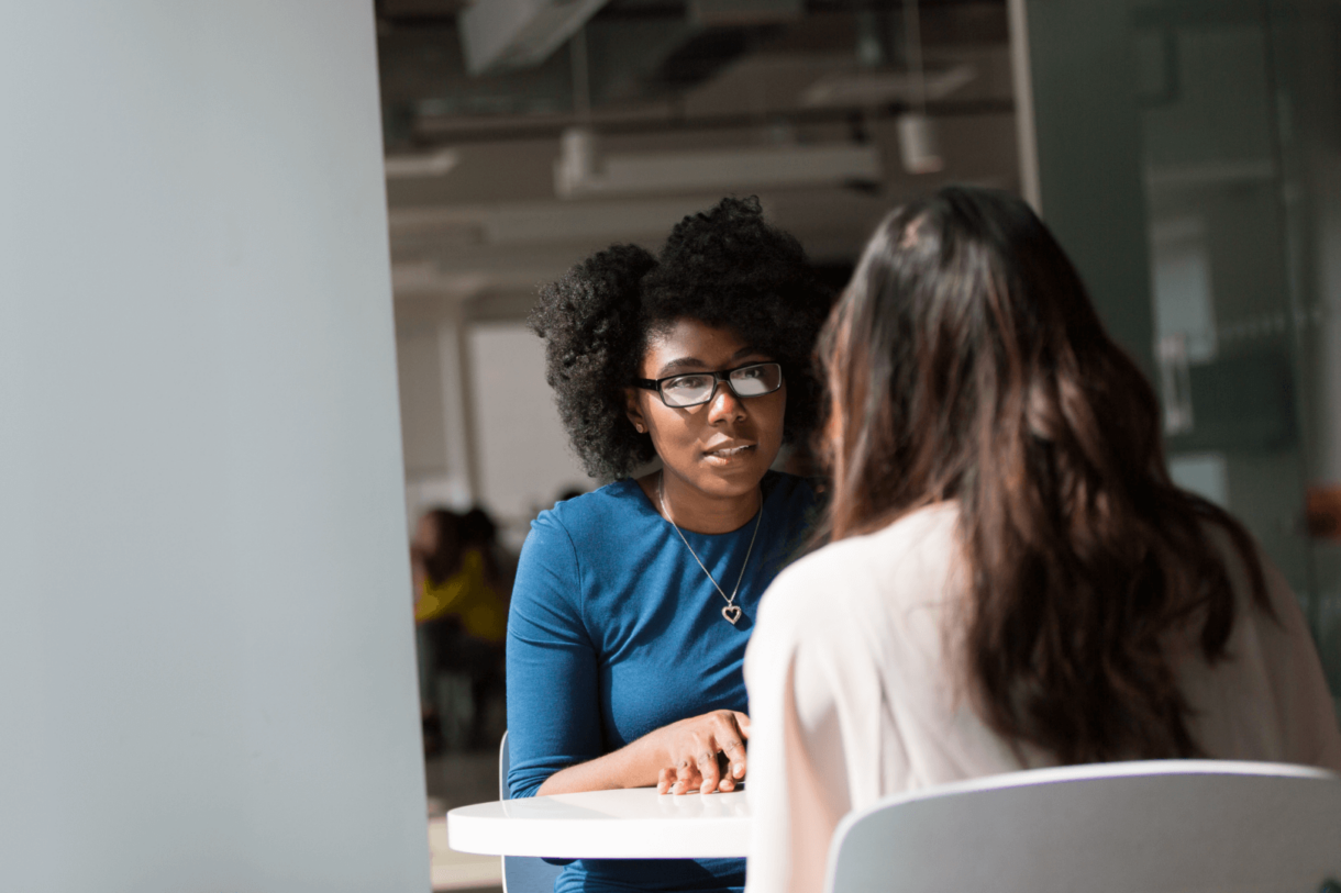 Two women sitting at a table having a conversation in a modern office setting.